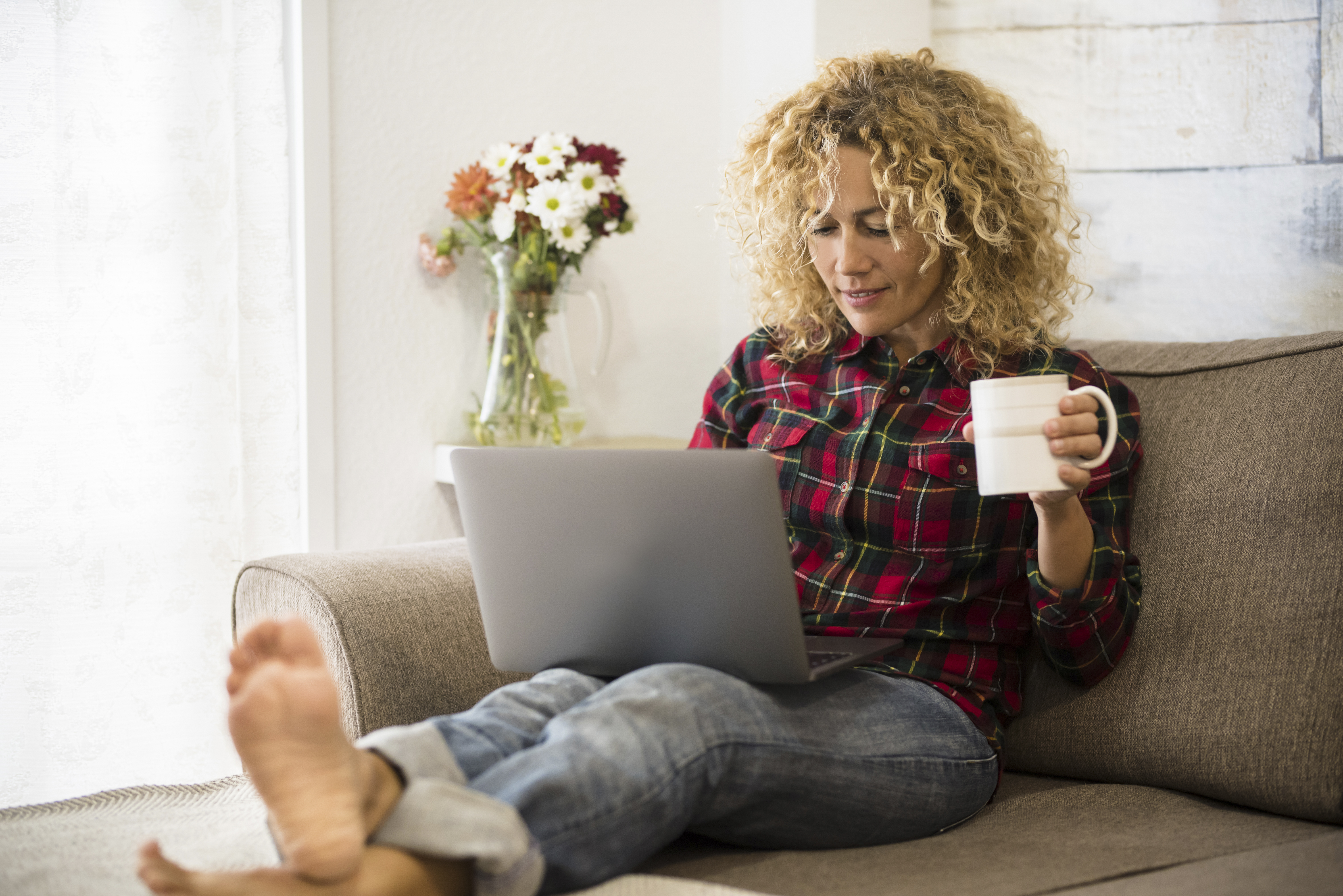 Image of a woman with her feet up on the coffee table, writing on her laptop and drinking tea