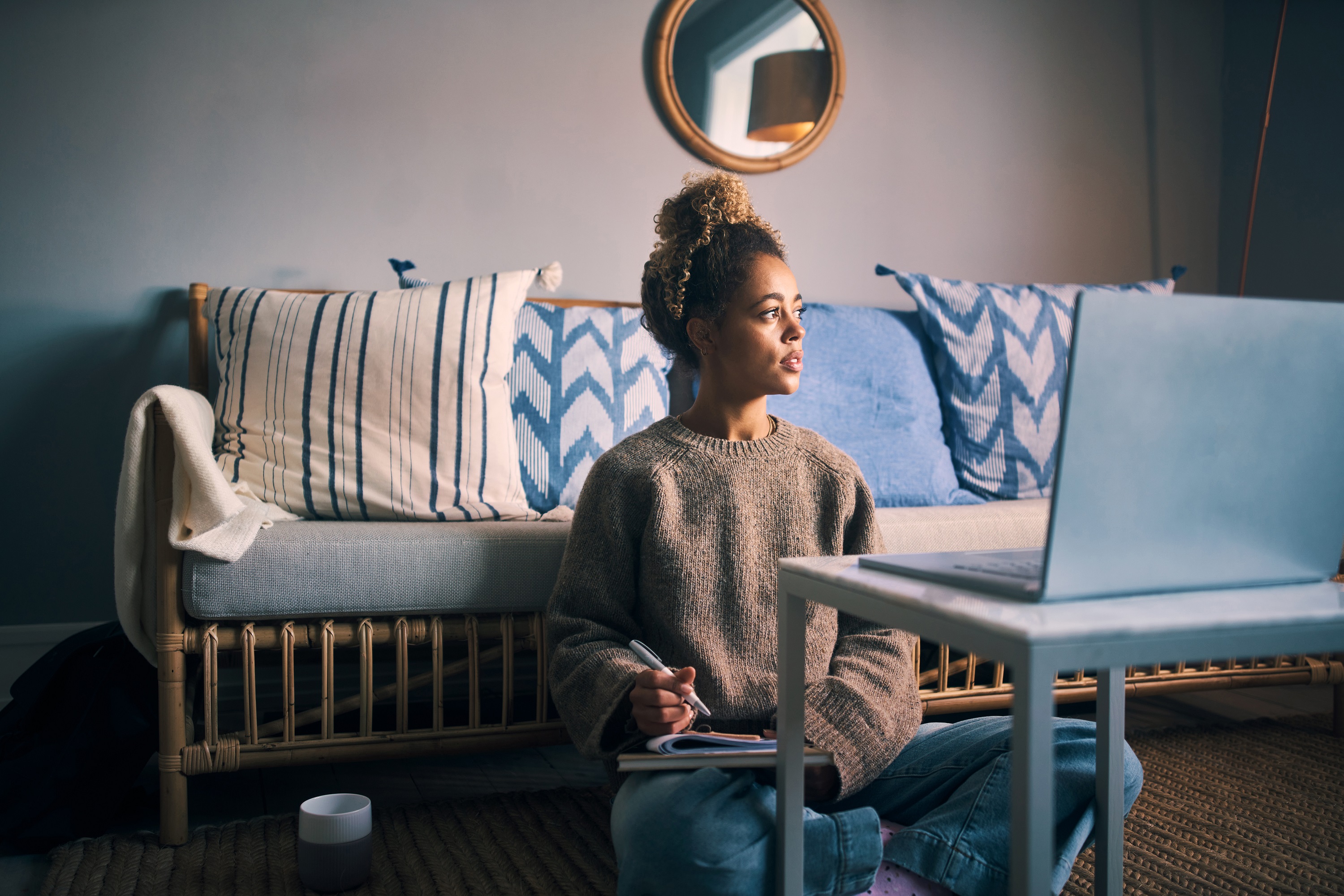 Woman sitting cross-legged on living room floor, thoughtful, with her laptop in front of her