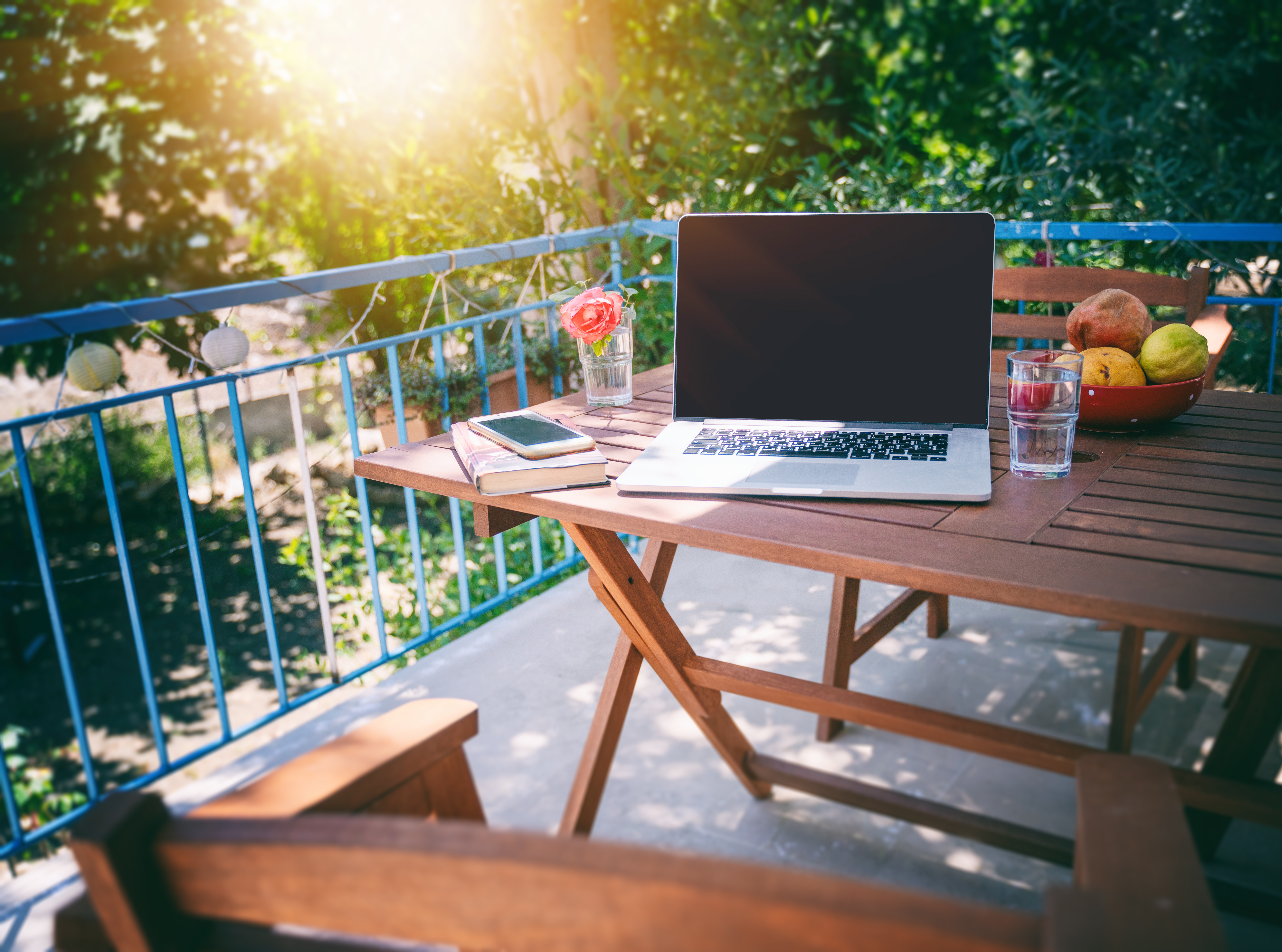 Image of a laptop left on a picnic table on a sunny terrace with blue railings
