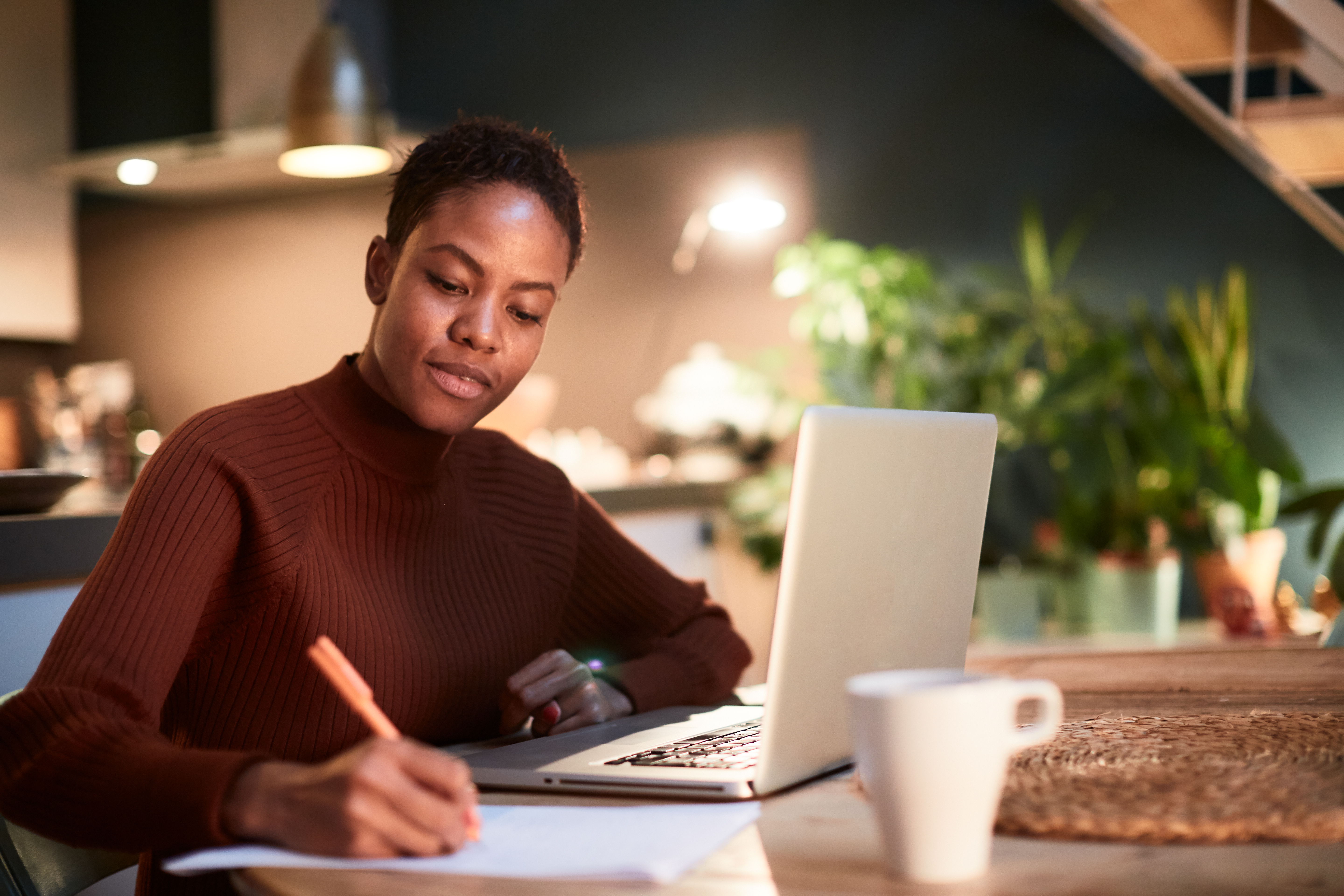 Image of a woman at large kitchen table, writing notes on a pad of paper with her laptop open in front of her.