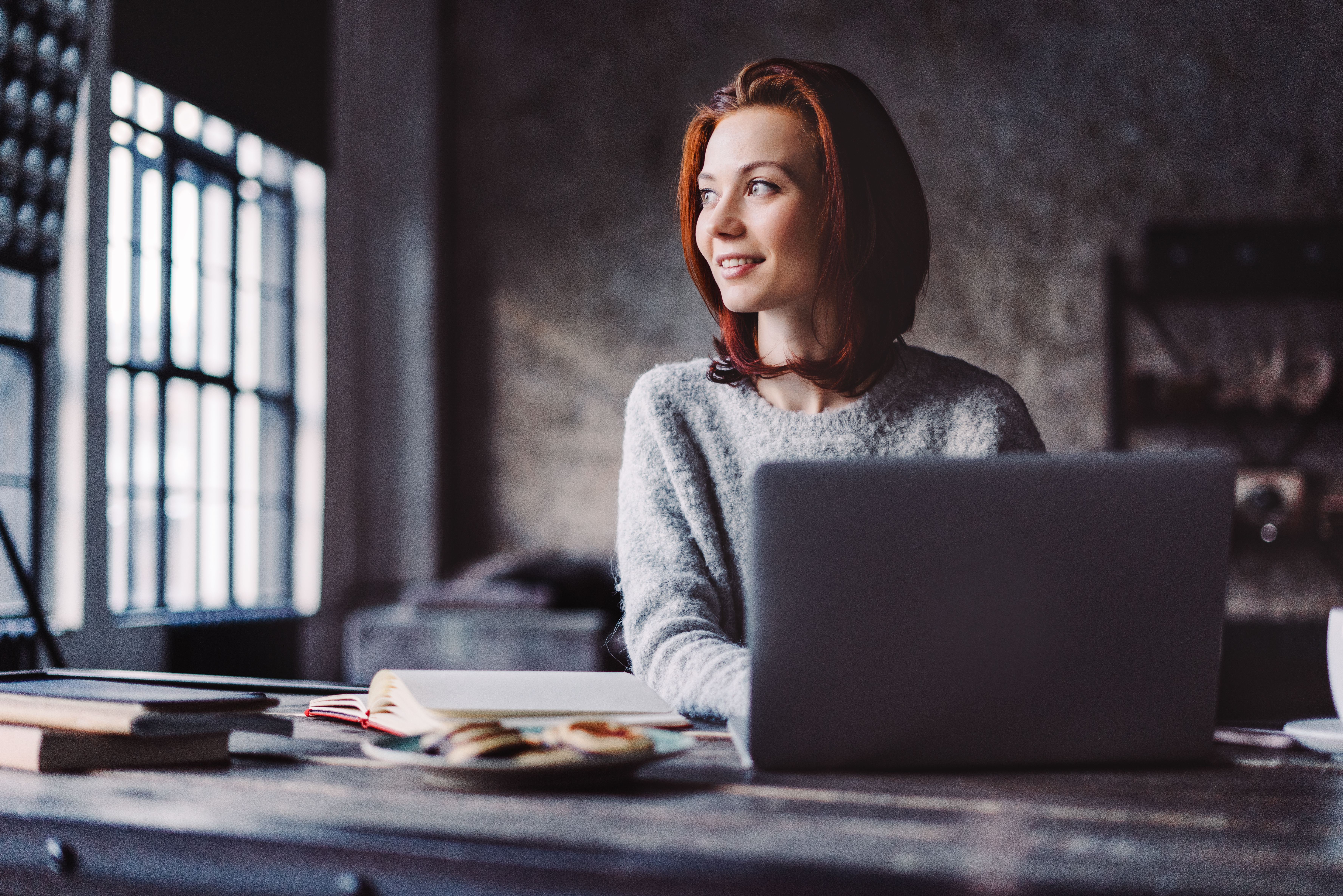 Image of a woman looking out of the window and smiling as she writes on her laptop